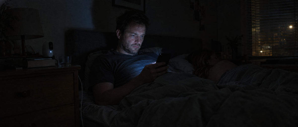 A man sits up in bed at 2am, uneasy, reading his phone while his partner sleeps